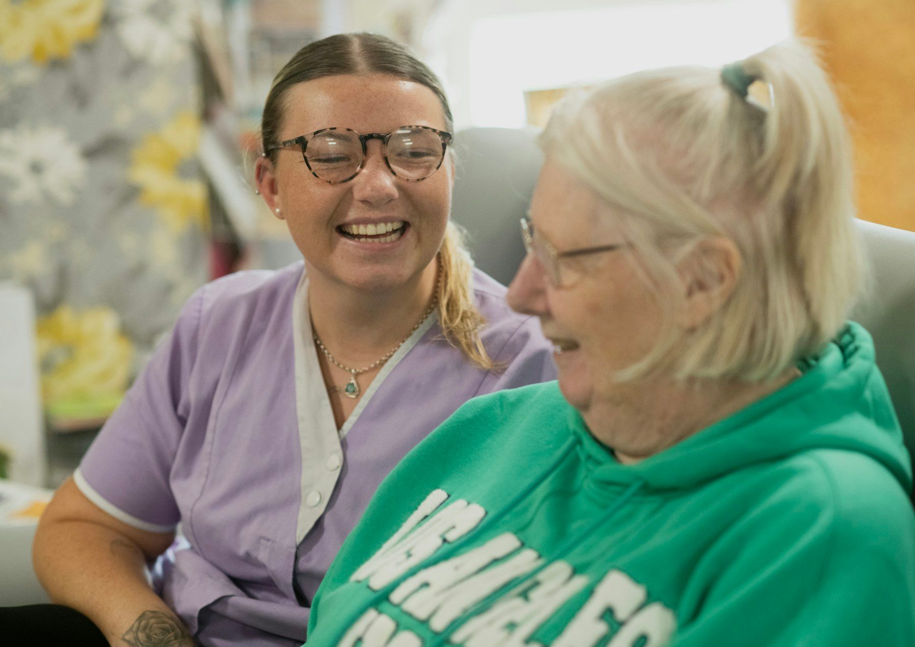 A young care worker and an older woman share a laugh while sitting together on a sofa.